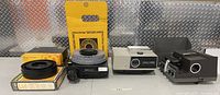 Photo showing three slide projectors from left to right: Airequipte 4400EF (black), Optina 500II (gray), Kodak 670H (black) with multiple spare slide carousels beside them.