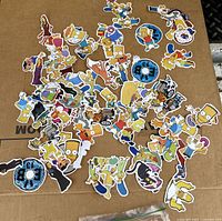 Top view of approximately 60 colorful character and themed stickers from The Simpsons laid out on a cardboard surface.