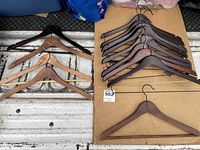 First image showing a collection of various wooden clothes hangers laid out on cardboard and a metal surface. Both natural and dark stained wood finishes visible, some hangers include trouser bars and hooks.
