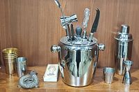 Overview of stainless steel bar set items arranged on wooden surface including ice bucket, shaker, strainer, cups, and tools.