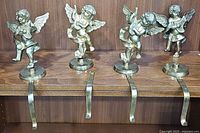 Four brass cherub stocking holders arranged in a row on wooden shelf, showing overall condition and design.