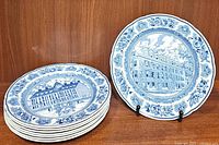 Seven blue and white Wedgwood plates stacked with one displayed upright showing intricate university building design and floral border.