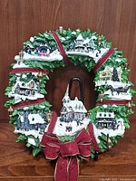Full front view of the ceramic Christmas wreath showing multiple miniature village houses, snow, greenery, and red ribbon with bow.