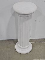 Full view of tall white Greek column pedestal standing against white wall and concrete floor, showing complete shape and size.