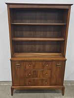 Front view of wooden mid-century hutch showing two shelves above and three drawers plus two doors below, with brass hardware.