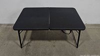 Front view of the black portable foldable table with legs extended, showing the full rectangular surface divided into two foldable halves.