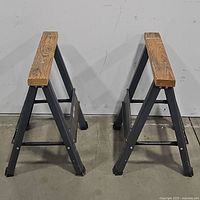 Pair of folding saw horses standing side by side, showing metal legs and wooden tops.