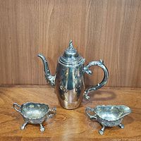Full view of the silver tea set including lidded tea pot, creamer, and sugar dish on a wooden surface.