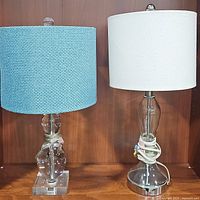Two table lamps side by side: acrylic base with blue textured shade on left and glass base with tan linen shade on right, shown from front.