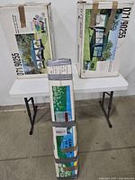 Three cardboard boxes displayed on a white folding table. The boxes contain a large canopy tent set with screen shades as indicated by the photos and text on the boxes.