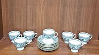 View of 8 Wedgwood Florentine tea cups arranged around 7 matching saucers stacked in the center on a wooden surface.