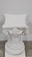 Front view of white ornamental pedestal planter stand showing carved floral and scroll design and fluted column shaft with visible damage on the top corner.