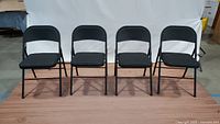 Front view of four identical black folding chairs with metal frames and upholstered seats in a row on wooden floor.