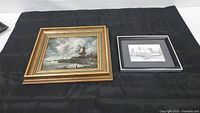 Two boat themed framed art pieces on black fabric: oil painting with windmill on left and watercolor with boats on right