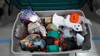 Large plastic tote filled with various Halloween decoration and craft items, including ghost figure, jack-o'-lantern buckets, small green and black decorative houses, spider webs with spiders, fabric Halloween bags, and colorful spirit hand decoration.