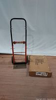 Metal hand truck with black handle and orange frame, two wheels visible, next to a sealed cardboard box containing the new dolly.
