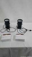 Two black metal spot lamps with cords and two white boxes on white surface.