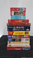 Stacked view showing twelve sealed puzzle boxes, piece counts 500-1000, multiple brands.