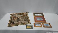 Overview of woven tapestry with rural village scene and five framed art prints arranged on table.