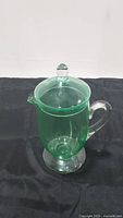 Green uranium glass pitcher with clear lid and handle sitting on a black fabric background.