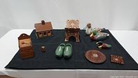 Nine assorted wooden decorative items arranged on black cloth including a painted log cabin, duck decoys, wooden clogs, carved plaques and a wooden calendar block.