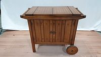 Front view of wooden bar cart showing closed folding top and cabinet doors with metal handles and wooden wheels on one side.