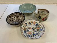 Overview of four pottery items: octagonal blue-green bowl, black and white patterned bowl, floral scalloped plate, and floral decorated teapot with woven handle.