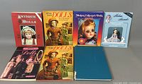 Seven collectible doll reference books arranged in two rows on a flat surface. Covers showing various antique and modern dolls.