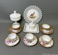 Full view of set showing milk glass trinket box, decorative plate with turkey, square plate with rabbit-topped trinket box, two purple and gold cup and saucer sets, and three floral porcelain cup and saucer sets.
