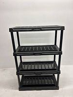 Front view of the black plastic four-shelf unit showing grid pattern on shelves and vertical supports.