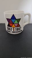 Front and side view of the coffee mug showing colorful star design with dates and text commemorating Canadian Confederation Centennial.