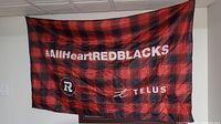 Red and black plaid 'All Heart Redblacks' flag with team and Telus logos hung on wall