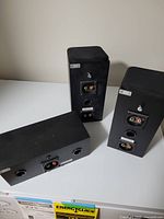 Photo showing the back of three black surround sound speakers with input terminals and bass reflex ports.