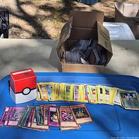 Overall view of trading cards spread on blue surface, Poké Ball box and additional cards in cardboard box