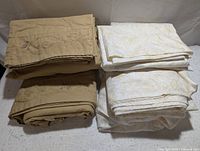 Photo showing folded beige and white bed sheet sets with embroidered and floral designs, includes flat, fitted sheets and pillowcases.