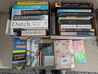 Image shows stacks of various books including fiction, travel guides, photography, and self-help. Bottom area shows three coloring books and a box of 24 colored pencils.