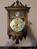 Front view of wooden chiming wall clock with brass face and winding key attached to the side.
