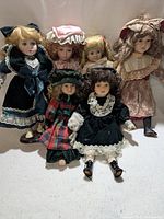 Six porcelain dolls displayed in a group with vintage Victorian-style clothing, lace details, and varied hair styles, some seated, some standing.