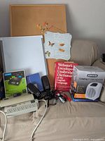 View of assorted office items including dictionary, heater in box, whiteboard, bulletin board, speakers, postal scale, cords, and other items arranged on a beige cloth-covered surface.