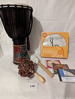 Photo showing the djembe drum with colorful patterned body, a small healing drum in an orange box, a wooden drumstick with white mallet head, a natural seed pod rattle, and multiple practice CDs.