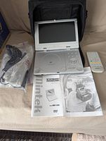 Portable DVD player with 8-inch TFT monitor, open and powered off, alongside remote control, power adapter, mounting strap, and manuals.