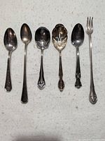 Top view of all six silver plated serving utensils arranged in a row on a white towel.