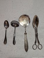 Top view of silver serving pieces including spoon, ladle, pie server, and tongs on a white background.