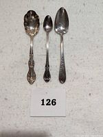 Three sterling silver spoons displayed side by side, showing front face with details of scalloped serving spoon, small spoon with floral handle, and plain spoon with engraved 'L'.