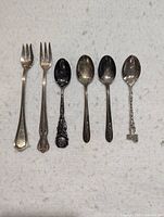 Top down photo of 2 forks and 4 spoons with tarnish and detailed designs, laid out side by side on a white cloth.