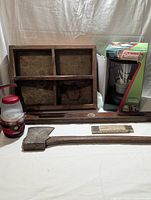 Two lanterns, wooden level, hatchet, folding measure, yardstick, mallet, and wooden box arranged on white surface with background curtain.