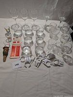 Full view of all bar items including multiple wine glasses, shot glasses, a figurine wine stopper, and white plastic plate clips spread out on a white surface.