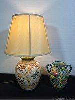 Lamp and vase shown side by side; lamp is hand-painted floral ceramic with beige shade, vase is green with raised floral design and two handles.