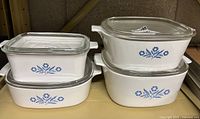 Four white ceramic CorningWare casserole dishes with blue cornflower motif and clear glass lids stacked in shelving.