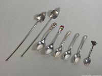 All eight spoons laid out, showing variety in size, handle design, and bowl shape, including enamel and elephant finial spoons.